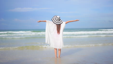 Back-of-Joyful-Woman-in-White-Tunic-on-Sandy-Beach-Raising-Arms-in-Front-of-Sea-Waves-and-Horizon
