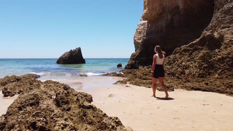 Playa-Praia-Dos-Tres-Irmaos,-Algarve,-Portugal---Niña-Camina-Por-La-Playa-De-Arena-A-Través-Del-Mar-Azul-Claro