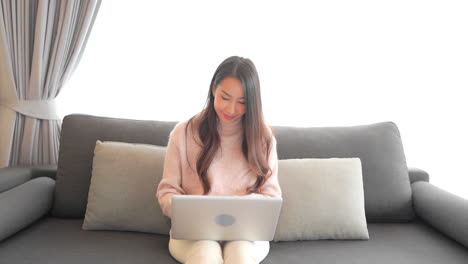 An-attractive-woman-in-a-sweater-and-white-jeans-sits-on-the-couch-while-she-works-on-her-laptop