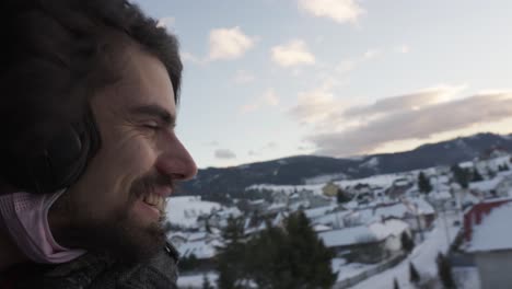 Bärtiger-Mann-Mit-Kopfhörern-Lächelnd-Mit-Kopf-Aus-Offenem-Fenster-Im-Zug-In-Verschneiter-Winterlandschaft,-Slowakei