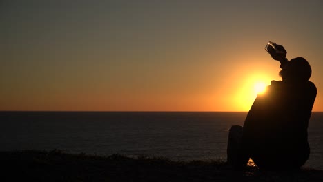 Silhouette-of-a-desperate-man-drinking-from-the-bottle-while-sitting-on-the-beach-at-sunset,-with-copyspace