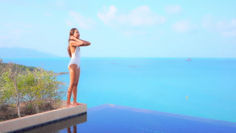 A-beautiful-sexy-woman-stands-on-the-edge-of-the-planter-in-a-resort-infinity-pool-while-looking-out-at-the-ocean-horizon