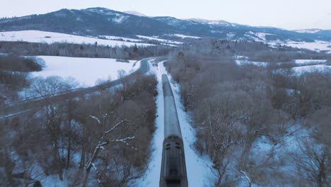 Incredible-aerial-drone-view-of-fast-moving-train-travelling-in-snowy-magical-white-landscape,-dusk,-circle-pan,-Slovakia-High-Tatra-winter-landscape