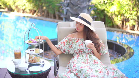 A-cute-woman-seated-on-a-poolside-lounge-chair-enjoys-a-sip-of-her-coffee-as-she-reaches-over-to-snag-a-sweet-breakfast-treat-from-the-food-caddy