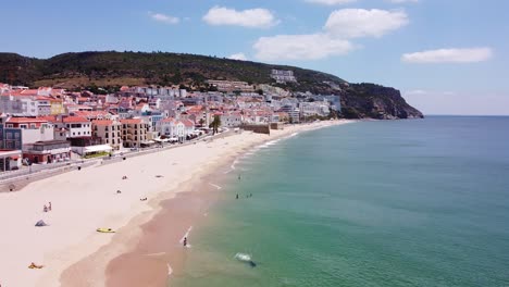 Praia-da-California-Beach-at-Sesimbra,-Alentejo,-Portugal---Aerial-Drone-View-of-the-Boulevard-with-Popular-Sandy-Beach,-Tourists,-Restaurants-and-Hotels