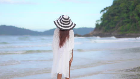 Back-of-Female-With-Floppy-Hat-and-Tunic-Walking-Alone-on-Beach-Sand-by-Tropical-Sea-Waves,-Slow-Motion