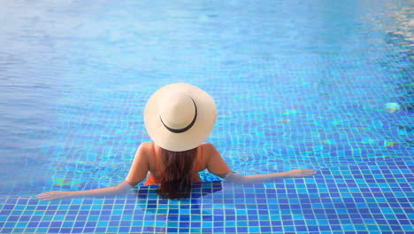 With-her-back-to-the-camera,-a-woman-wearing-a-straw-sunhat-lounges-in-a-swimming-pool