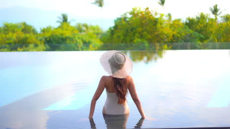 A-woman-with-her-back-to-the-camera-sits-on-the-shallow-step-of-a-swimming-pool-as-she-looks-out-at-the-tropical-landscape-and-ocean-horizon-beyond