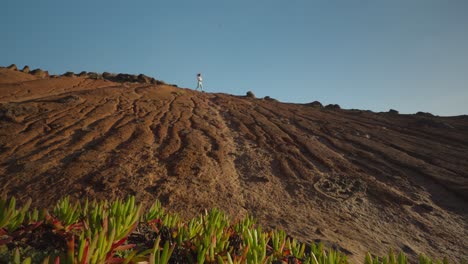 Woman-on-orange-sand-hill-walking-down,-green-vegetation-in-foreground,-hike-adventure