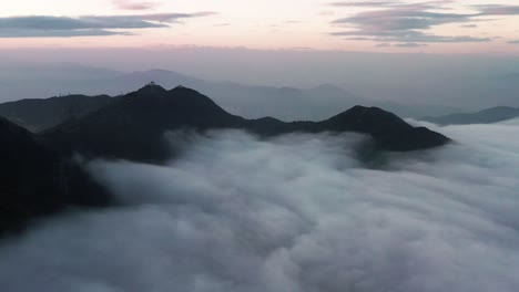 Aerial-dolly-in-flying-over-dense-layer-of-clouds-covering-mountain-tops-at-blue-hour-in-Kowloon-Peak,-Hong-Kong,-China