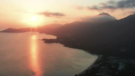 Aerial-truck-left-of-sea-and-Pui-O-beach-bay-near-village-and-rainforest-hills-at-golden-hour-with-sunbeams,-Lantau-Island,-Hong-Kong,-China