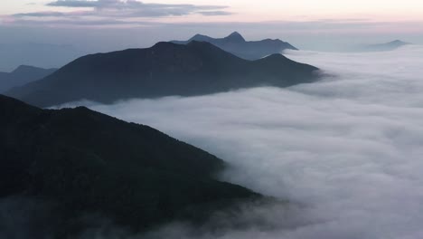 Aerial-parallax-of-dense-layer-of-clouds-passing-between-mountain-tops-at-blue-hour-in-Kowloon-Peak,-Hong-Kong,-China