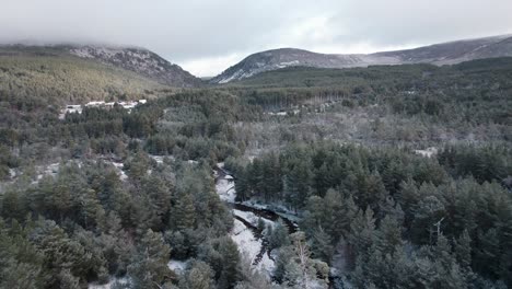 Cinematic-drone-footage-flying-towards-a-mountain-landscape-above-the-snow-covered-canopy-of-a-Scots-pine-forest-and-frozen,-icy-river