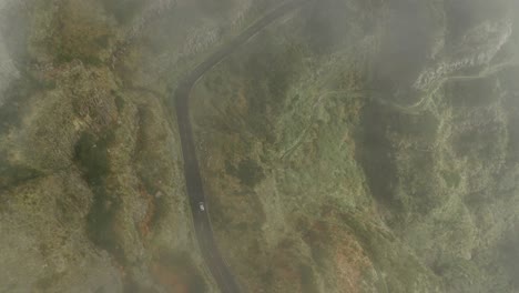 Above-car-driving-through-rugged-mountain-terrain,-reveal-shot-through-mist,-aerial