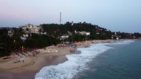 Aerial-Drone-Over-Sandy-Beach-Dining-Tourist-Destination-Busy-With-People,-Dogs-And-Umbrellas-At-Sunset-In-Sri-Lanka