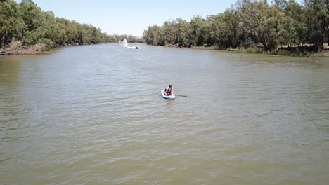 Drohnenantenne-über-Fluss-Frau-Kniet-Auf-Stand-Up-Paddle-Board-Boot-Fährt-Mit-Wasserskifahrer-Vorbei
