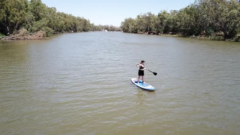 Drone-aerial-moving-backwards-over-river-woman-standing-up-on-paddle-board