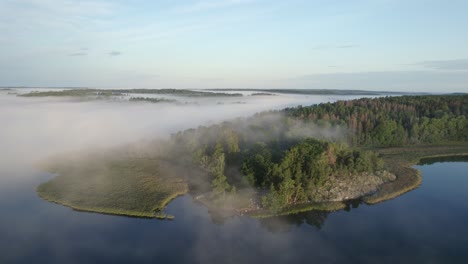 Morning-sun-on-Swedish-wooded-lake-headland-partially-obscured-by-mist,-drone-orbit