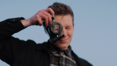 Young-caucasian-male-photographer-taking-picture-with-analog-photo-camera-during-sunset-against-the-blue-sky