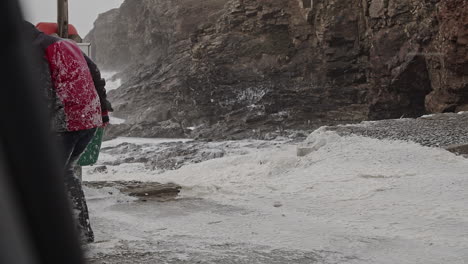Woman-Covered-In-Snow-like-Sea-Foam-During-Extreme-Winter-Storm-Eunice-At-The-Coast-Of-Chapel-Porth-Beach-In-Cornwall,-UK