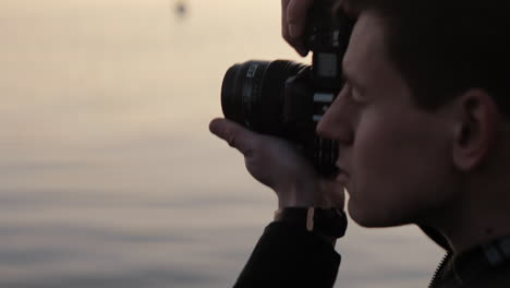 Young-caucasian-photographer-taking-a-picture-with-an-analog-camera-at-a-lake-sunset-focus