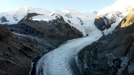 Aerial-flyover-across-the-end-of-Morteratsch-glacier-in-Engadin,-Switzerland-at-dawn-with-a-pan-down-from-some-of-the-tallest-peaks-of-the-Swiss-Alps-like-Piz-Bernina,-Piz-Palu