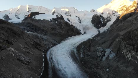 Aerial-flyover-over-the-end-of-Morteratsch-glacier-in-Engadin,-Switzerland-at-dawn-with-a-pan-down-from-some-of-the-tallest-peaks-of-the-Swiss-Alps-like-Piz-Bernina,-Piz-Palu
