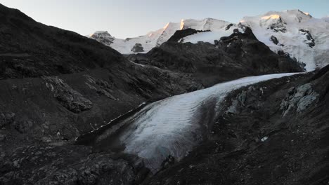 Aerial-flyover-towards-the-end-of-Morteratsch-glacier-in-Engadin,-Switzerland-at-dawn-with-a-view-of-from-some-of-the-tallest-peaks-of-the-Swiss-Alps-like-Piz-Bernina,-Piz-Palu