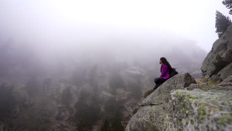 Solo-traveller-young-woman-backpacker-sitting-alone-on-the-edge-of-Rocky-Mountain-cliff-with-foggy-natural-landscape