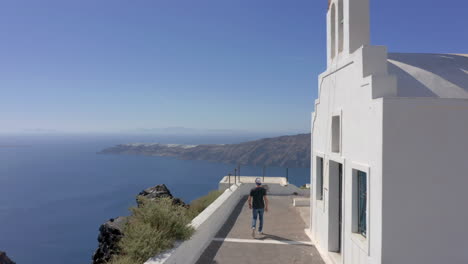 Aerial:-One-man-walks-next-to-a-church-in-Santorini,-Greece-on-a-sunny-day