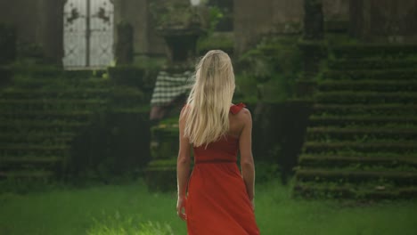 Mysterious-blond-woman-in-red-dress-walking-on-grass-field-towards-stone-steps-of-ancient-temple,-fantasy-scene