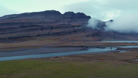 Ovejas-Pastando-En-Un-Campo-De-Hierba-Junto-A-Un-Río-Debajo-De-Un-Macizo-Rocoso,-Islandia