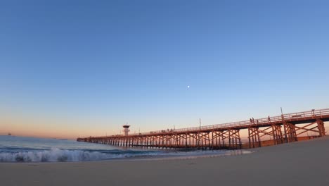 The-moon-setting-over-the-beach-with-a-long-wooden-pier-at-sunrise---seascape-time-lapse