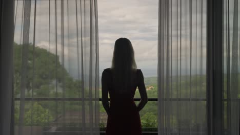 Silhouette-of-woman-waking-up-and-stepping-outside-of-cabin-house-balcony,-curtains-slowly-swaying-in-wind