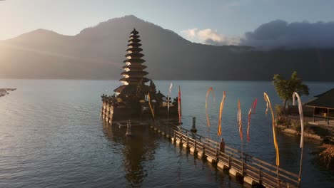 Woman-walks-towards-Hindu-temple-on-Lake-Batur-with-light-rays-from-sunrise,-mountain-background,-holy-morning,-aerial