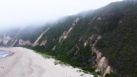 Aerial-drone-shot-of-Sedgefield-beach-surrounded-by-fertile-sand-dunes-covered-in-mist-from-low-hanging-clouds,-below-people-go-about-their-day-playing-on-the-sand,-Western-Cape,-South-Africa