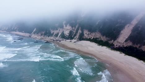 Aerial-drone-shot-descending-below-a-blanket-of-clouds-panning-to-reveal-a-deserted-beach-on-a-miserable-day,-Sedgefield,-Western-Cape,-South-Africa
