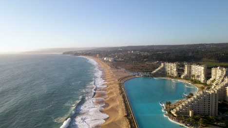 Flying-over-a-huge-swimming-pool-next-to-modern-buildings-at-beach-of-Algarrobo,-Chile