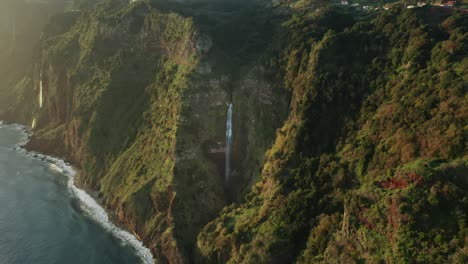 Sunrise-illuminating-grand-coast-line-of-Madeira-with-waterfall,-golden-hour,-aerial