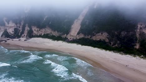 Aerial-drone-shot-hovering-below-the-mist-of-low-hanging-clouds-which-are-pushing-up-into-the-sand-dunes-that-surround-the-beautiful-secluded-beach-in-Sedgefield,-Western-Cape,-South-Africa