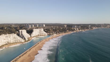 Flying-over-a-huge-swimming-pool-next-to-modern-buildings-at-beach-of-Algarrobo,-Chile
