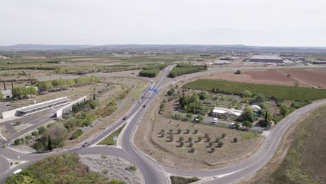 Traffic-circle-roundabout-with-cars-on-their-way-to-entering-town-of-Clermont-l'Hérault-France,-Aerial-approach-shot