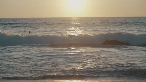 Eine-Große-Welle-Aus-Dem-Pazifischen-Ozean-Bricht-Auf-Die-Untiefen-Eines-Sandstrandes-In-Mexiko,-Während-Mehrere-Personen-Während-Des-Sonnenuntergangs-Schwimmen