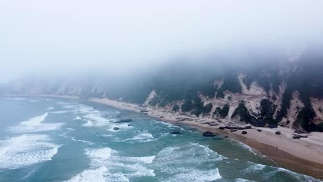An-epic-aerial-drone-shot-flying-through-low-hanging-clouds-along-a-choppy-Indian-Ocean-shoreline-in-Sedgefield,-Western-Cape,-South-Africa
