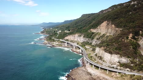 Aerial:-Cars-driving-along-a-curving-highway-next-to-a-stunning-ocean-coastline,-in-New-South-Wales-Australia