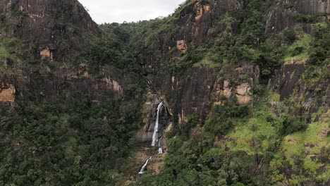 Toma-Aérea-Lejana-De-Una-Cascada-Profunda-Que-Fluye-Suavemente-Entre-Enormes-Montañas-Verdes,-Sri-Lanka