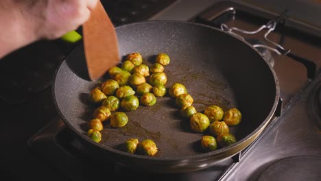 A-close-up-shot-of-marinated-brussels-sprouts-roasting-on-a-gas-hob-in-a-kitchen,-as-a-chef-carefully-tosses-and-stirs-the-vegetables-with-a-wooden-spatula-to-ensure-an-even-cook-on-a-hot-frying-pan