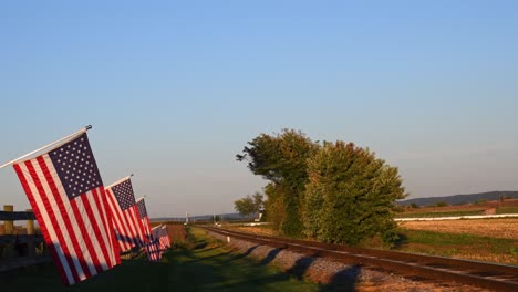 Ein-Blick-Auf-Eine-Linie-Sanft-Wehender-Amerikanischer-Flagge-Auf-Einem-Zaun-Von-Ackerland-An-Einem-Einzigen-Eisenbahngleis-Am-Späten-Nachmittag