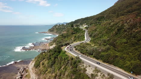 Aerial:-Drone-flying-over-vehicles-driving-along-a-highway-next-to-a-beautiful-ocean-coastline,-in-New-South-Wales-Australia