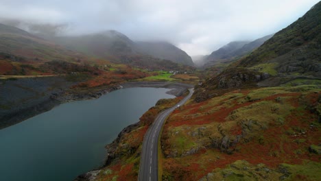 Car-driving-on-road-along-lake-of-Dinorwic-quarry-with-mountains-shrouded-in-clouds-in-background,-Wales-in-UK